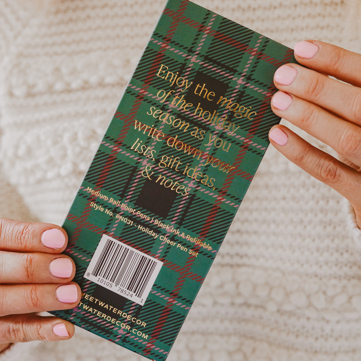 A person holds a green plaid holiday card with gold text that says, 'Enjoy the magic of the holiday season as you write down your list, gift ideas & notes.' The background shows a cozy, textured cream knit sweater.