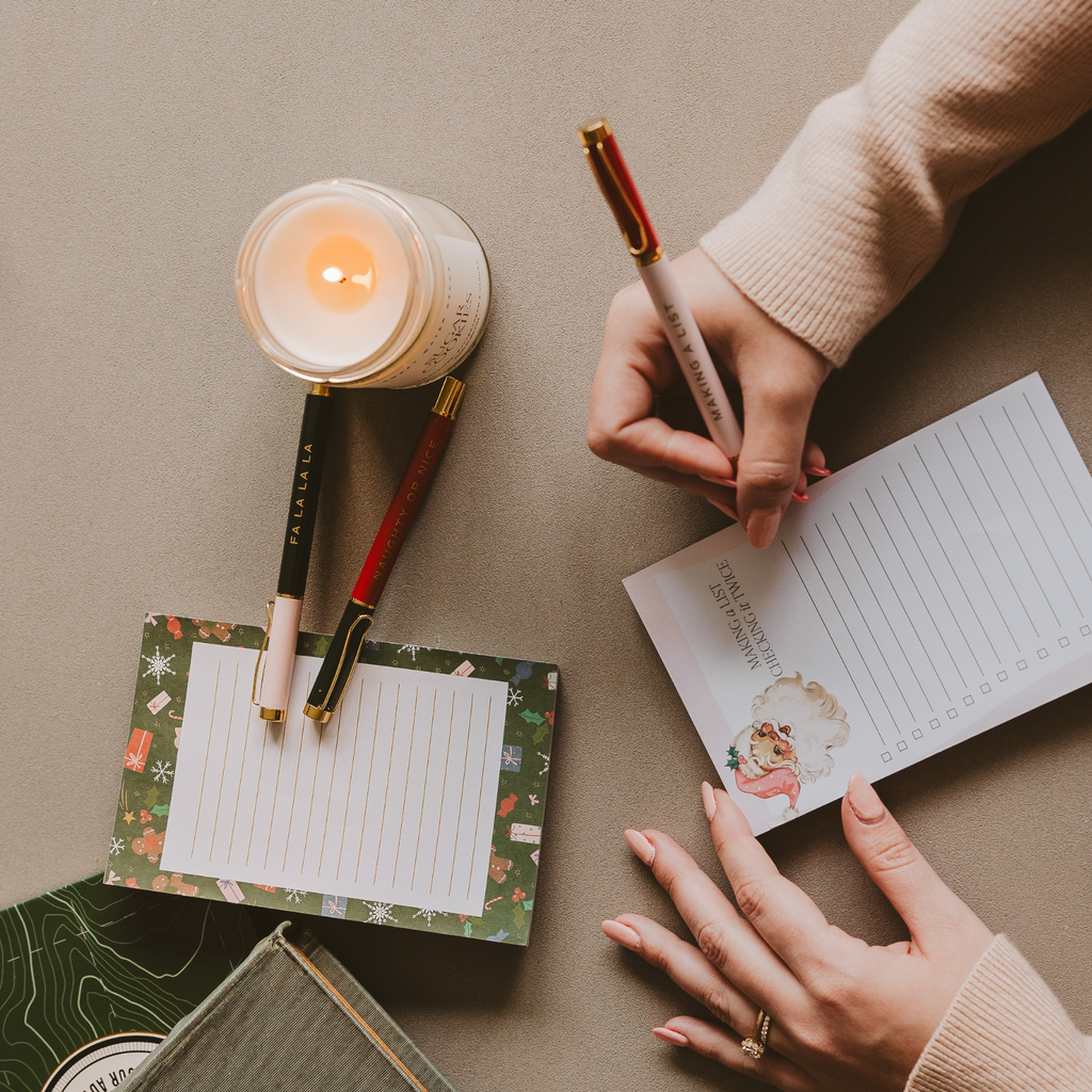 Person holding a pen over a notepad with a lit candle and small notepads on a table.