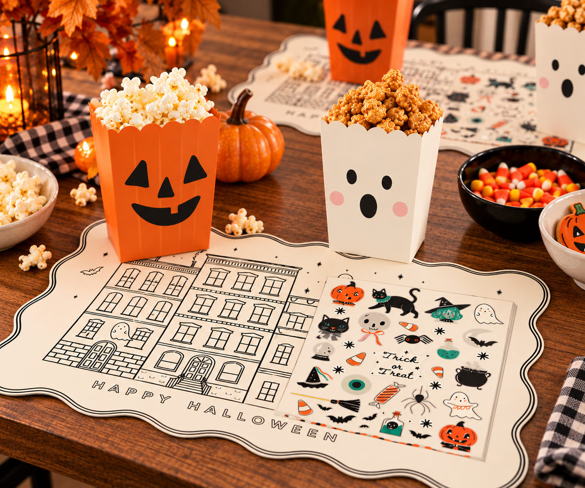 A Halloween-themed tabletop scene with printed placemats, popcorn boxes, candy bowls, pumpkins, and fall decor arranged on a wooden table.
