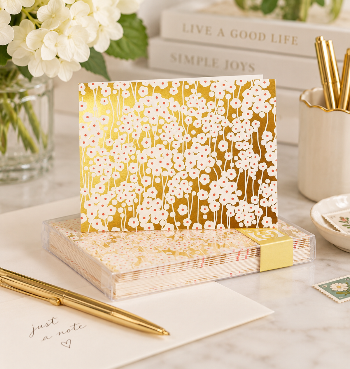 A stack of floral note cards with a shiny gold cover featuring white cherry blossom patterns, displayed on a marble desk with a gold pen, books, and flowers.